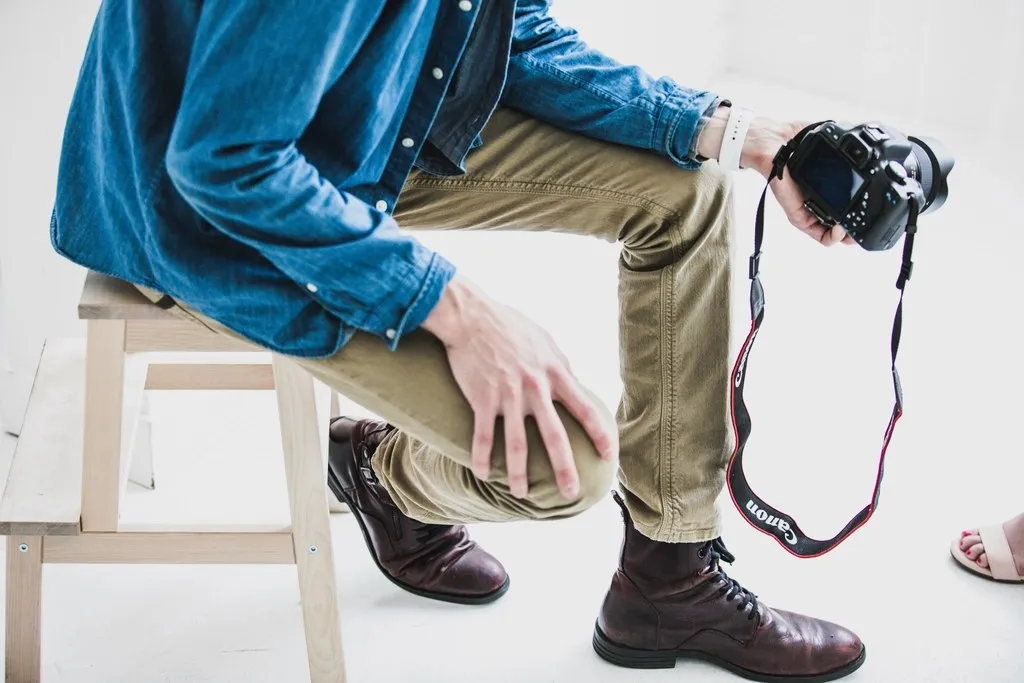 Muškarac u platnenim pantalonama i tamnim kožnim cipelama do članka sedi na stolici sa fotoaparatom u ruci